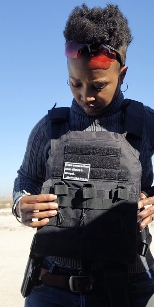 A woman standing outside in a desert area is removing a black tactical vest with a patch on the front, all over a gray turtleneck sweater. She is wearing blue jeans with a black holster attached. She has red-lensed shooting glasses on her up and resting on her forehead.