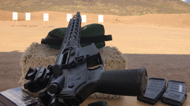 AR rifle on its side pointed downrange with action open. Two empty magazines on the shooting table to the right. An unopened box of ammunition to the left.